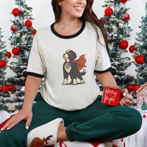 May include: Cream t-shirt with black trim, featuring a superhero dog graphic. The dog wears a red cape and mask, with an American flag design on its chest. A red mug with "Merry & Bright" is held.