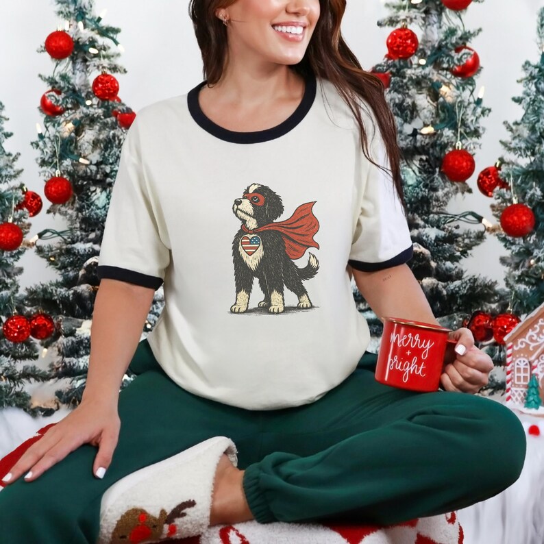 May include: Cream t-shirt with black trim, featuring a superhero dog graphic. The dog wears a red cape and mask, with an American flag design on its chest. A red mug with "Merry & Bright" is held.