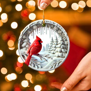 Peut inclure: Ornement de No&euml;l rond et blanc repr&eacute;sentant un cardinal rouge perch&eacute; sur une branche enneig&eacute;e. L'ornement pr&eacute;sente une sc&egrave;ne hivernale avec des arbres enneig&eacute;s et des flocons de neige. Il est suspendu par une ficelle dor&eacute;e.