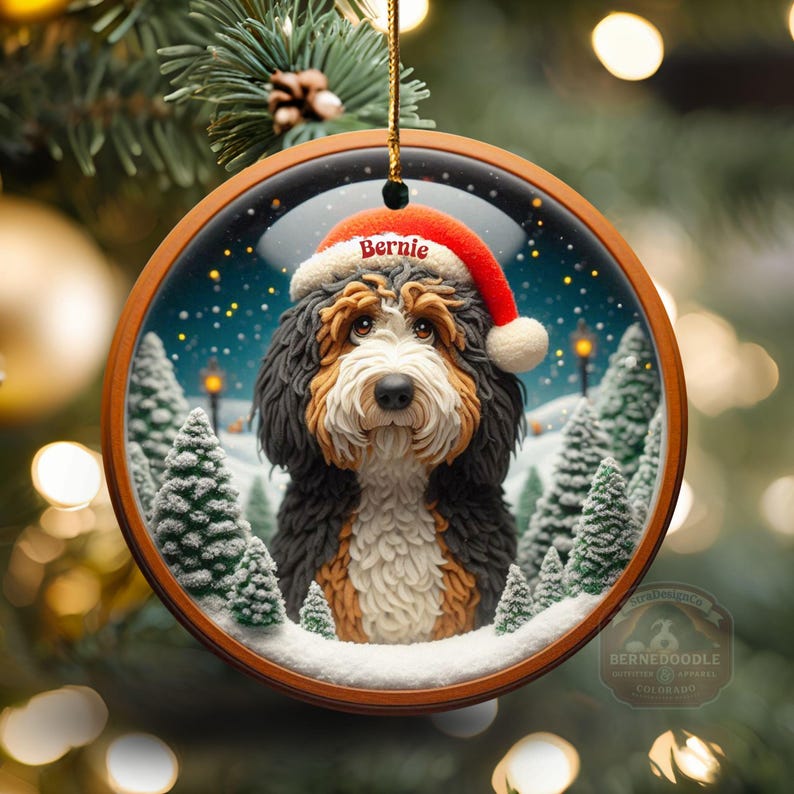 Op de afbeelding: Ronde kerstversiering met een Bernedoodle hond met een kerstmuts met de naam "Bernie". De versiering heeft een houten frame en een winterse achtergrond met besneeuwde bomen en een sterrenhemel. Het hangt aan een gouden touwtje.