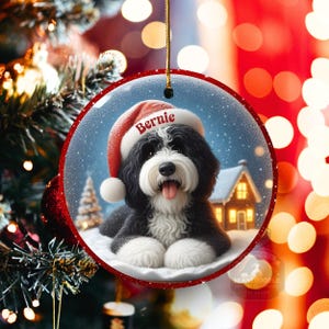 May include: A round Christmas ornament featuring a black and white Bernedoodle dog wearing a Santa hat with the name "Bernie". The ornament has a snowy winter scene with a house and Christmas tree. The ornament is trimmed with red.