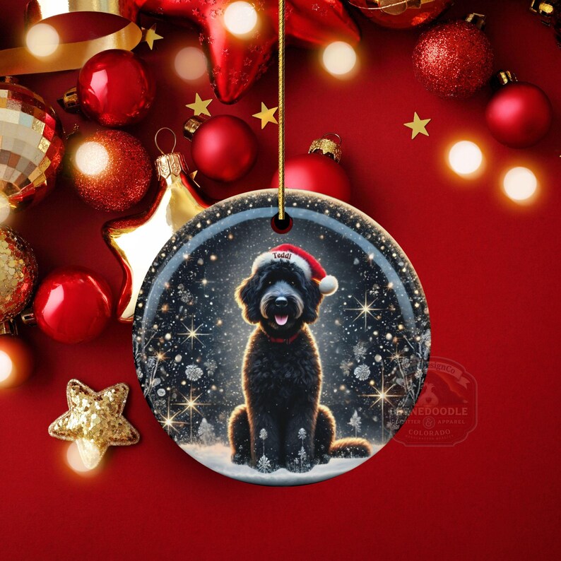 May include: A round Christmas ornament featuring a black dog wearing a Santa hat, set against a snowy winter scene. The ornament has a gold string for hanging and is surrounded by red and gold holiday decorations.