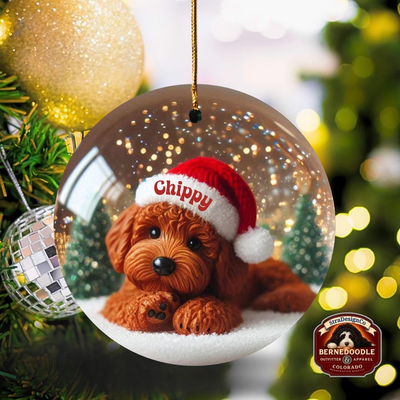 May include: A clear, round Christmas bauble featuring a brown dog wearing a Santa hat with the name "Chippy" in red. The bauble has a gold string for hanging and is set against a festive background with a Christmas tree and lights.