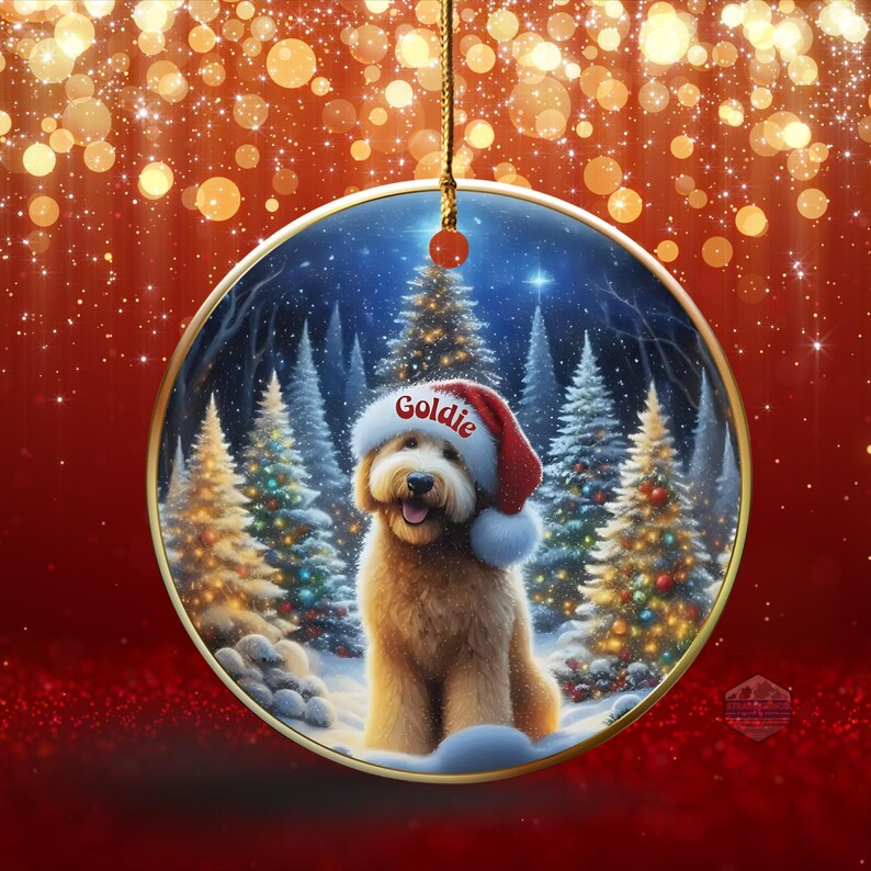 May include: A round Christmas ornament featuring a golden retriever wearing a Santa hat with the name "Goldie" on it. The dog is set against a snowy forest scene with Christmas trees. The ornament hangs from a gold string.