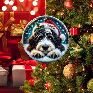 May include: A round Christmas ornament featuring a black and white dog wearing a Santa hat with the name "Bernie". The ornament has a snowy background with holly and stars. The ornament is hanging on a Christmas tree.