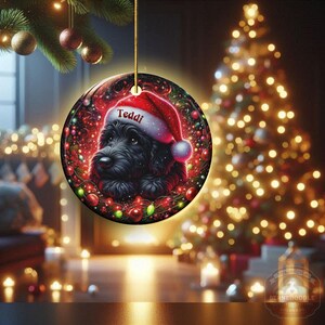 May include: A round Christmas ornament featuring a black dog wearing a Santa hat with the name "Teddi" on it. The ornament has a festive design with red and green accents, and is hanging in front of a lit Christmas tree.