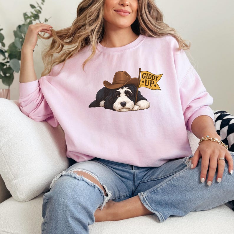 Puede incluir: Sudadera rosa claro con un perro de dibujos animados con sombrero de vaquero y una bandera con la frase "Giddy Up". El perro es blanco y negro. La sudadera es de color liso.