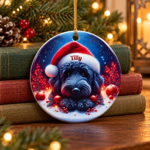 May include: A round ceramic Christmas ornament featuring a black dog wearing a red and white Santa hat. The name Tilly is printed in red on the hat brim. The background shows a snowy scene with red berries and glowing red ornaments.