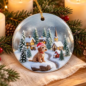 May include: A round ceramic Christmas ornament featuring a golden-brown dog wearing a red Santa hat with the name Goldie printed on it. The scene depicts a snowy village with pine trees, small houses, and a church, all under a light snowfall.