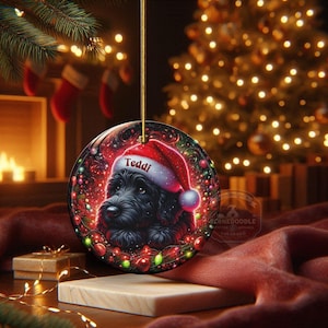 May include: A round Christmas ornament featuring a black dog wearing a Santa hat with the name "Teddi". The ornament has a red and green festive design with a gold string for hanging. The background includes a Christmas tree and a fireplace.