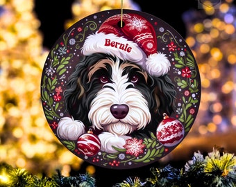 May include: A round Christmas ornament featuring a dog wearing a Santa hat with the name "Bernie". The ornament has a wreath of leaves and red berries around the dog's face, with two red and white ornaments.