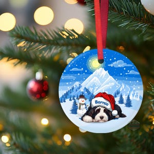 May include: Round Christmas ornament with a winter scene featuring a black and white dog wearing a Santa hat that says "Bernie". The ornament has a red ribbon for hanging and a snowy mountain backdrop.
