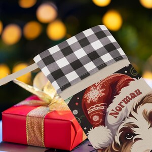 A festive Christmas stocking featuring a whimsical illustrated doodle dog wearing a red Santa hat, surrounded by holiday ornaments and greenery in a warm, detailed Christmas art style. The top of the stocking has a classic red and black buffalo-plaid cuff, and the stocking hangs from a white mantel decorated with evergreen garland and gold ornaments. The stocking design is personalized with your dog’s name, making it a charming and custom holiday décor piece for dog lovers.