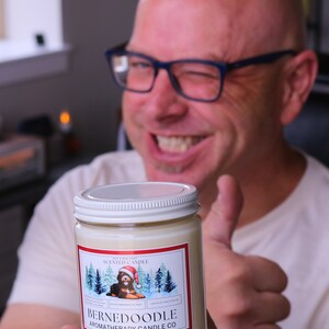 Puede incluir: Vela arom&aacute;tica en un tarro de vidrio con tapa blanca. La etiqueta presenta un perro Bernedoodle con un gorro de Pap&aacute; Noel en una escena invernal. La vela es de Bernedoodle Aromatherapy Candle Co.