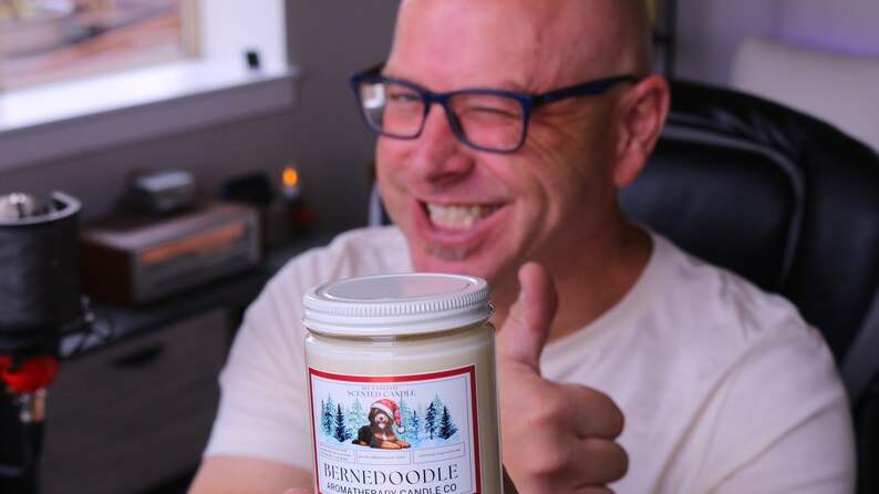 Puede incluir: Vela arom&aacute;tica en un tarro de vidrio con tapa blanca. La etiqueta presenta un perro Bernedoodle con un gorro de Pap&aacute; Noel en una escena invernal. La vela es de Bernedoodle Aromatherapy Candle Co.