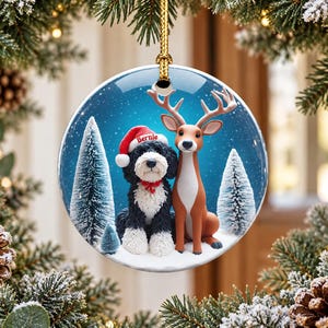 May include: A round ceramic Christmas ornament featuring a black and white dog wearing a red Santa hat with the name Bernie printed on it, sitting next to a brown deer in a snowy forest with pine trees against a blue background.