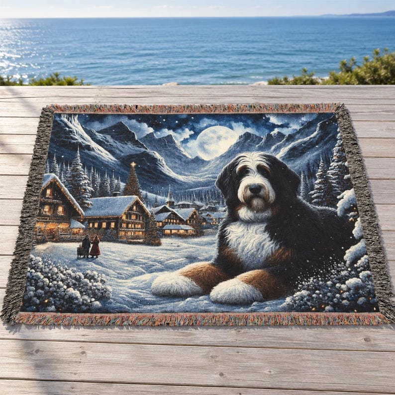 Puede incluir: Una manta tejida que muestra a un perro de monta&ntilde;a de Berna tumbado en primer plano de un pueblo nevado. El fondo muestra monta&ntilde;as cubiertas de nieve, pinos y una luna llena bajo un cielo nocturno oscuro y estrellado.