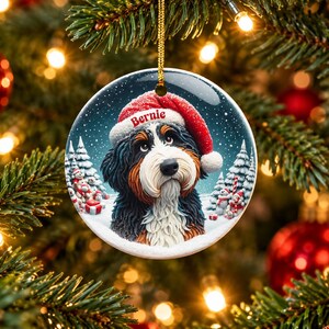 May include: A round ceramic Christmas ornament featuring a Bernese Mountain Dog wearing a red Santa hat with the name Bernie printed on the brim. The dog is set against a snowy winter landscape with pine trees and small snowmen.