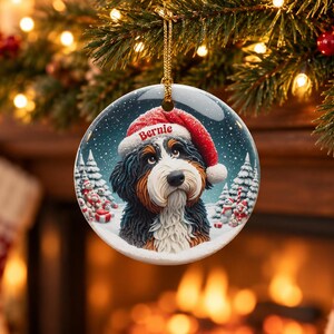 May include: A round ceramic Christmas ornament featuring a Bernese Mountain Dog wearing a red Santa hat with the name Bernie printed on the brim. The background shows a snowy winter landscape with pine trees and small snowmen figures.