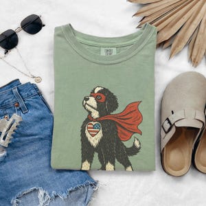 May include: A sage green crewneck t-shirt featuring a graphic of a black and white dog wearing a red superhero mask and cape, with a heart-shaped American flag patch on its chest. The inner neck label displays the text Comfort Colors.