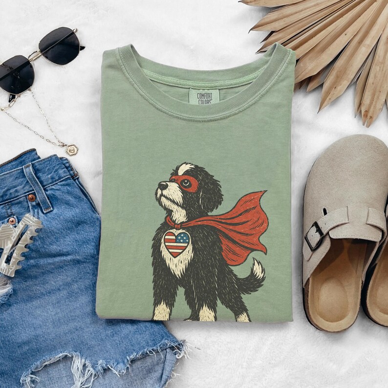 May include: A sage green crewneck t-shirt featuring a graphic of a black and white dog wearing a red superhero mask and cape, with a heart-shaped American flag patch on its chest. The inner neck label displays the text Comfort Colors.
