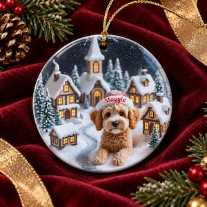 May include: A round ceramic Christmas ornament featuring a fluffy golden-brown dog wearing a white Santa hat with the name Maggie printed in red. The background depicts a snowy village scene with houses, a church, and pine trees.