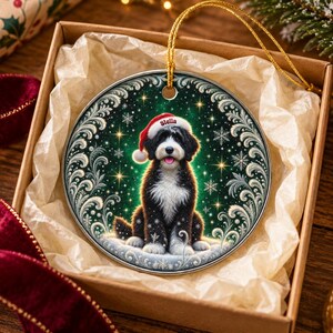 May include: A round ceramic ornament featuring a black and white dog wearing a red Santa hat with the name Stella printed on it. The dog sits against a dark green background with white snowflake patterns and a decorative silver border.