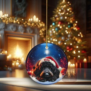 May include: A round Christmas ornament featuring a black dog wearing a Santa hat with the name "Stella". The ornament has a blue background with a star and snow, with a Christmas tree and fireplace in the background.