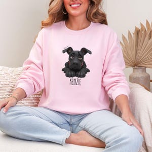 Puede incluir: Sudadera rosa claro con un dise&ntilde;o de perro negro y el nombre "KENZIE" debajo. El perro tiene una expresi&oacute;n triste. La sudadera tiene cuello redondo y mangas largas, ideal para un look casual.