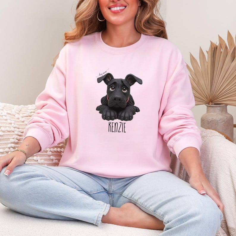 Puede incluir: Sudadera rosa claro con un dise&ntilde;o de perro negro y el nombre "KENZIE" debajo. El perro tiene una expresi&oacute;n triste. La sudadera tiene cuello redondo y mangas largas, ideal para un look casual.