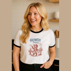 May include: White ringer t-shirt with a black collar and sleeves. The shirt features a graphic of a cowboy dog with the text "HOWDY DOODLE" in blue. The dog is holding a lasso and standing near cacti.
