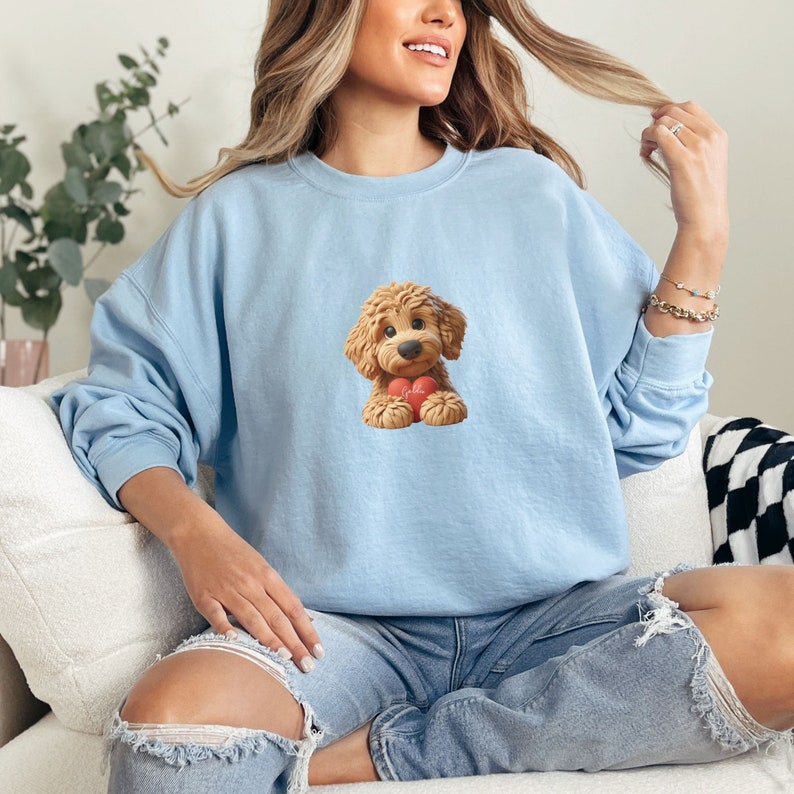 May include: A light blue sweatshirt featuring a cartoon illustration of a fluffy, brown dog holding a red heart with the word "Hello" on it. The sweatshirt has long sleeves and a crew neck.