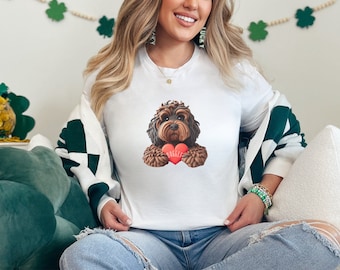 Peut inclure: T-shirt blanc avec un chien marron et noir tenant un cœur rouge avec le mot "Hugger". Le chien a une apparence duveteuse. La personne porte un pull blanc et vert et un jean bleu.