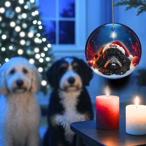 May include: A round Christmas ornament featuring a dog wearing a Santa hat, with the name "Stella" on it. The ornament hangs in front of a Christmas tree and two dogs. Two lit candles are in the foreground.