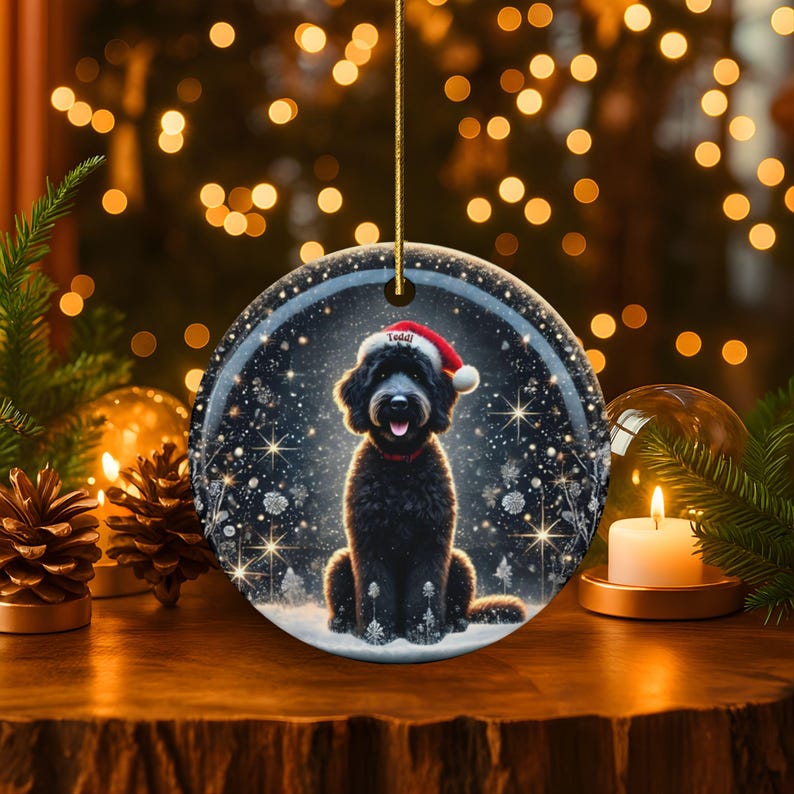 May include: A round Christmas ornament featuring a black dog wearing a Santa hat with the name "Teddy" on it. The ornament has a winter scene with snowflakes and trees. The background has blurred lights and greenery.