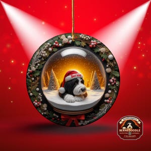 May include: A round Christmas ornament featuring a snow globe design with a black and white dog wearing a Santa hat that says "Bernie". The ornament is surrounded by a wreath and has a red bow.