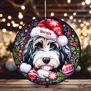 以下が含まれることがあります： 「バーニー」という名前のサンタ帽をかぶった犬が描かれた丸いクリスマスオーナメント。オーナメントは、暗い紫色の背景に花のボーダーと赤と白のクリスマスオーナメントが付いています。犬は黒、白、茶色の毛皮をしています。
