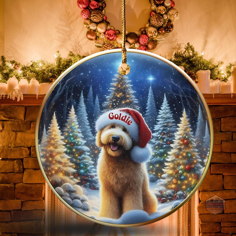 May include: A round Christmas ornament featuring a golden doodle dog wearing a Santa hat with the name "Goldie" on it. The background shows a snowy forest with Christmas trees. The ornament is hanging in front of a fireplace.