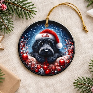 May include: A round ceramic Christmas ornament featuring a black dog wearing a red Santa hat with the name Stella printed on the brim. The dog is surrounded by red berries, shiny red ornaments, and a snowy blue background with a gold string.