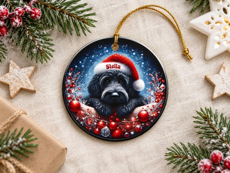 May include: A round ceramic Christmas ornament featuring a black dog wearing a red Santa hat with the name Stella printed on the brim. The dog is surrounded by red berries, shiny red ornaments, and a snowy blue background with a gold string.