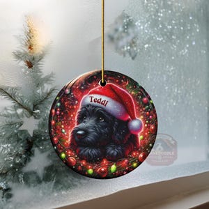 May include: A round Christmas ornament featuring a black dog wearing a Santa hat with the name "Teddi" on it. The ornament is surrounded by festive red and green lights and a red background. A gold string is attached for hanging.