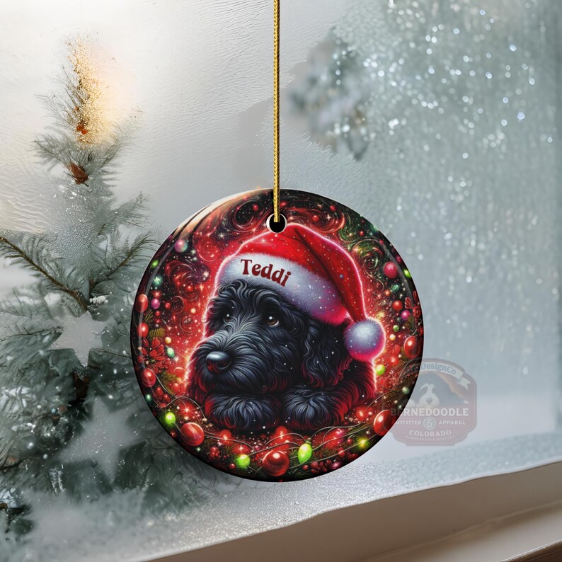 May include: A round Christmas ornament featuring a black dog wearing a Santa hat with the name "Teddi" on it. The ornament is surrounded by festive red and green lights and a red background. A gold string is attached for hanging.