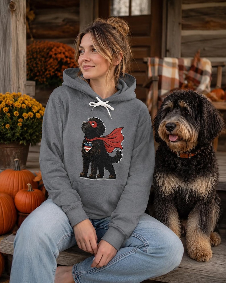 Pu&ograve; includere: Una felpa con cappuccio grigio melange con la grafica di un cane nero che indossa una maschera e un mantello da supereroe rossi, con un'etichetta a forma di cuore con motivo a stelle rosso, bianco e blu sul petto e cordini bianchi.