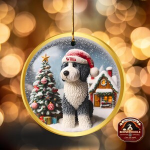 May include: A circular Christmas decoration featuring a Bernerdoodle dog wearing a Santa hat with the name "Willis". The ornament depicts a winter scene with a Christmas tree, a snow-covered house, and a gold border. It is suspended by a gold string.