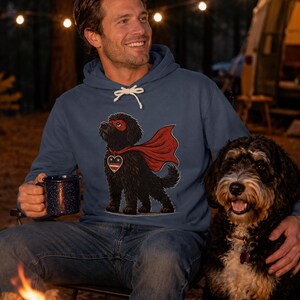 Pu&ograve; includere: Una felpa con cappuccio blu caratterizzata da un'illustrazione grafica di un cane nero che indossa un mantello da supereroe rosso, una mascherina rossa e un emblema a forma di cuore sul petto con un motivo a stelle. La felpa ha lacci bianchi.