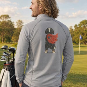 K&ouml;nnte beinhalten: Ein hellgrauer Langarm-Pullover mit kurzem Rei&szlig;verschluss, der auf der R&uuml;ckseite eine Grafik eines schwarz-wei&szlig;en Hundes zeigt, der einen roten Umhang mit einem herzf&ouml;rmigen amerikanischen Flaggendesign tr&auml;gt. Das Kleidungsst&uuml;ck hat Raglan&auml;rmel.