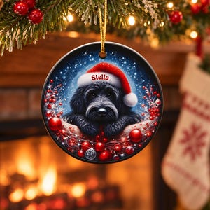 May include: A round ceramic Christmas ornament featuring a black dog wearing a red and white Santa hat with the name Stella printed on the brim. The dog is surrounded by red berries, red ornaments, and a snowy blue background.