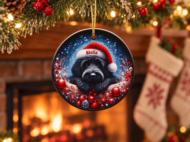 May include: A round ceramic Christmas ornament featuring a black dog wearing a red and white Santa hat with the name Stella printed on the brim. The dog is surrounded by red berries, red ornaments, and a snowy blue background.