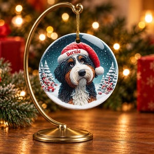 May include: A round ceramic ornament hanging from a gold metal stand. The ornament features a Bernese Mountain Dog wearing a red Santa hat with the name Bernie printed on the brim, set against a snowy winter landscape with pine trees.