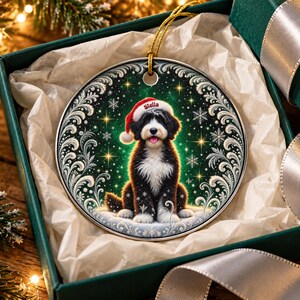 May include: A round ceramic Christmas ornament featuring a black and white dog wearing a red Santa hat with the name Stella printed on the brim. The dog is centered against a dark green background with white snowflakes and silver filigree.